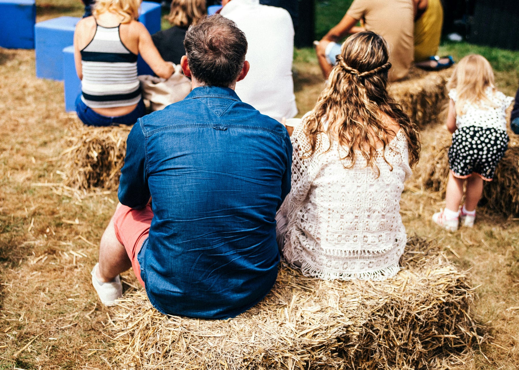 Couple sitting on a hay bale at a festival with children playing in background